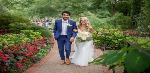 Couple walking on a brick pathway. A man is wearing a navy blue suit and white shirt and a woman is wearing a white wedding gown, holding ivory flowers.
