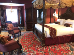 Romantic overnight guest room with red carpets, burgundy bed furnishings, fabric chairs and a king-size bed.