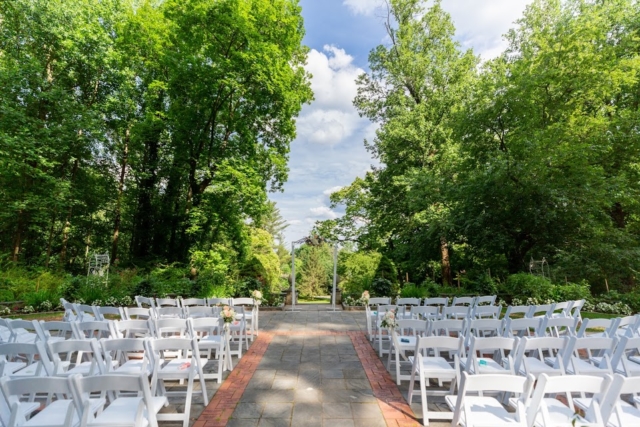 Outdoor ceremony setup with white chairs surrounded by green trees.