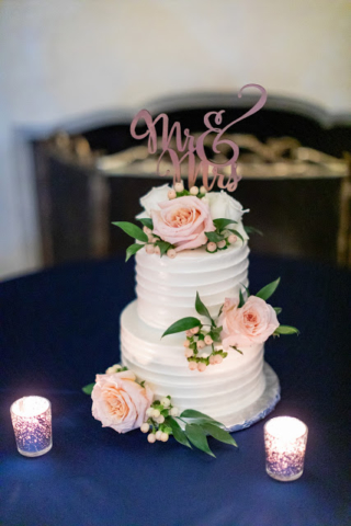 White wedding cake with pink roses and a pink cake topper. Candles are in front of the cake.
