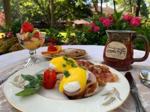 Eggs Benedict with bacon, a pepper. fresh fruit, blueberry pancakes and a maroon coffee mug.