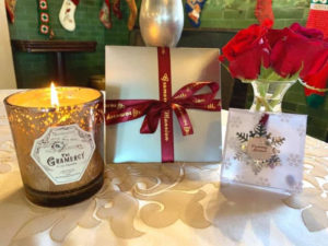 A lit candle in a brown glass holder, a gift certificate with red ribbon and gold lettering and a silver snowflake ornament sit in front of red roses on a table.